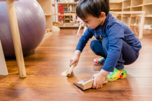 A young boy in a blue shirt holds a small brush and sweeps into a dustpan.