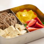 A metal bento box with soba noodles, tofu, and strawberries