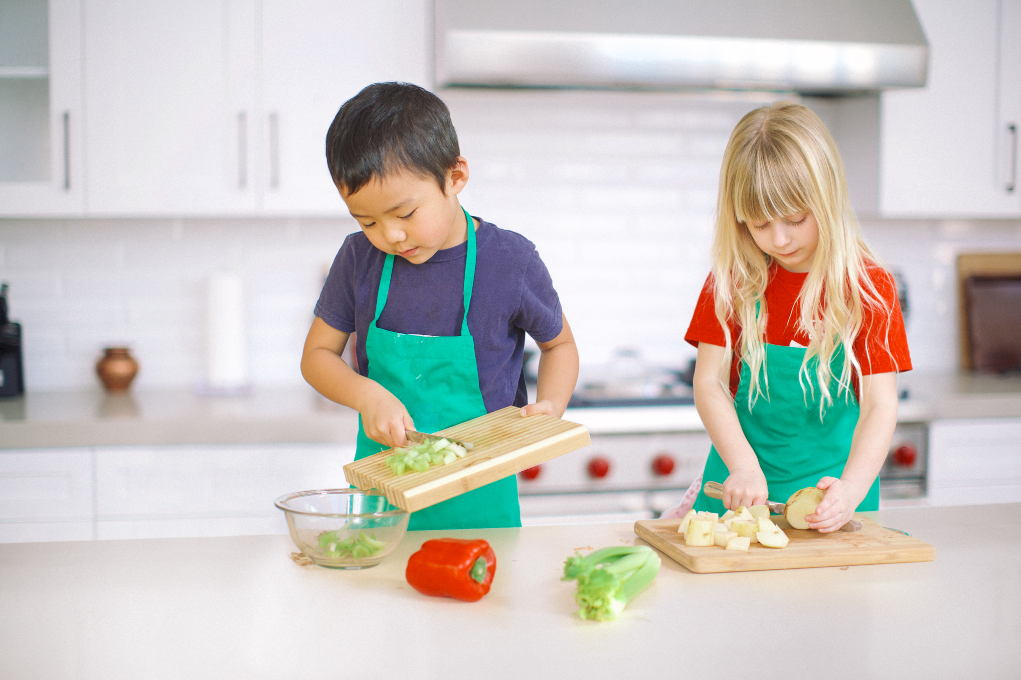 A girl and a boy wear teal aprons and cut vegetables in a kitchen